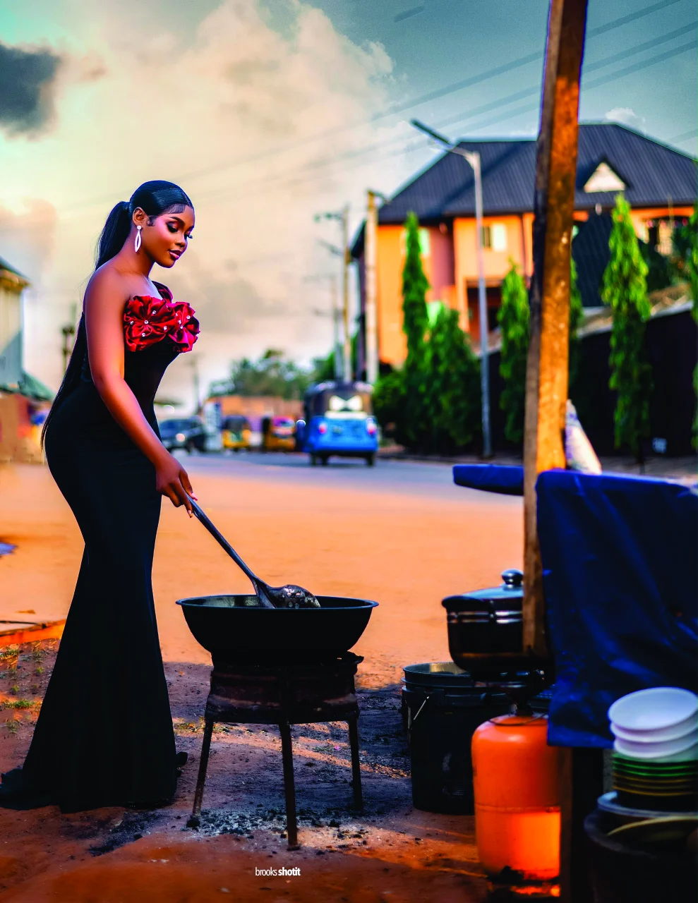 Chiamaka frying akara by the roadside at sunset in a black gown with red rosettes