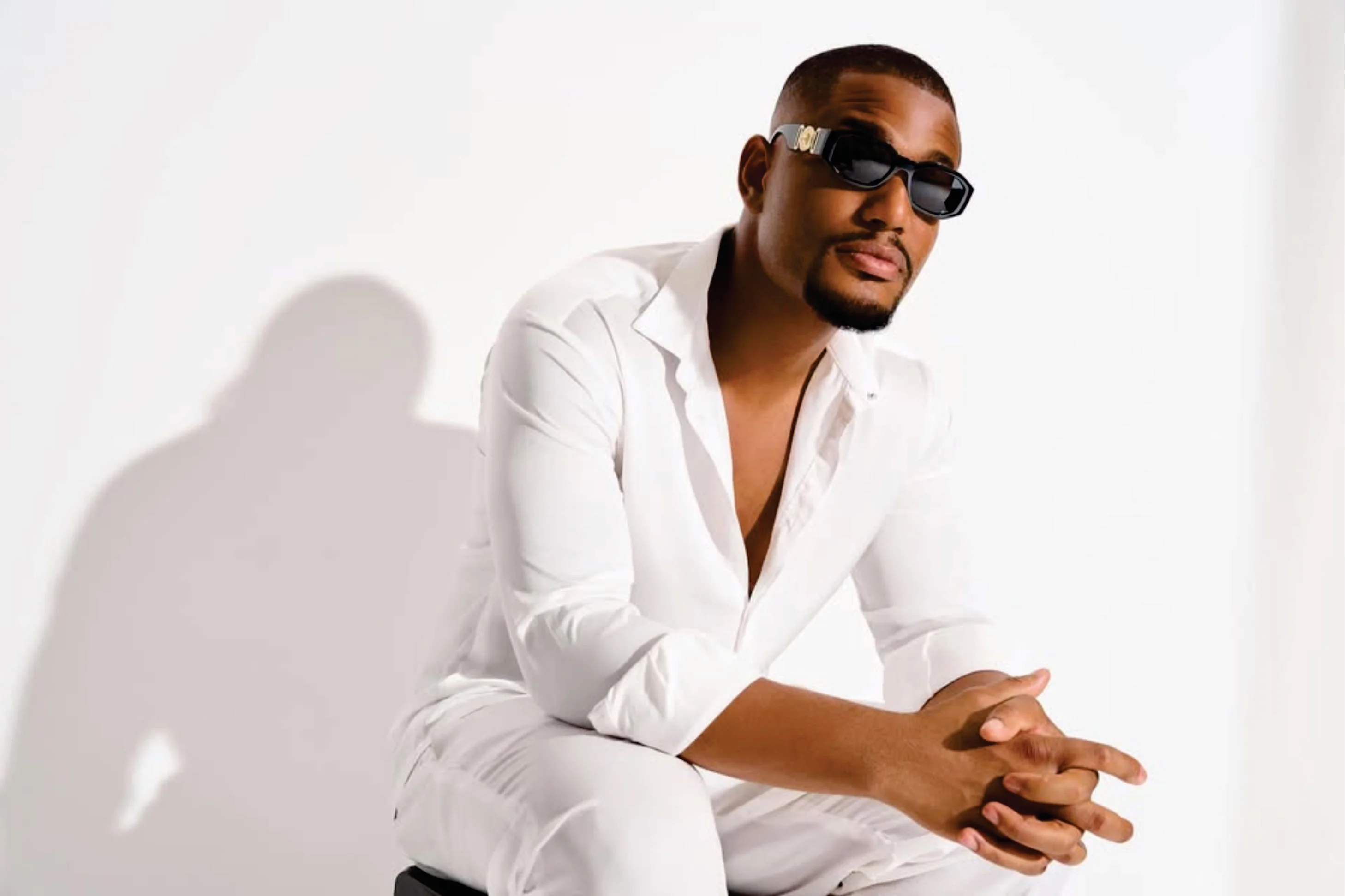 Alex Ekubo in a white suit and sunglasses, hands clasped